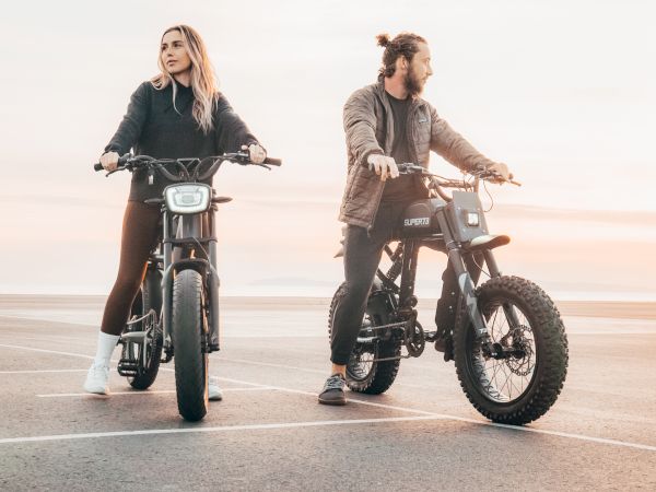 Two people are sitting on electric bikes in an open area, with a beautiful sunset in the background.